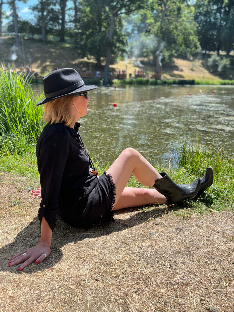 Girl in black dress and hat wearing black cowboy boot wellies sitting on the edge of a lake.