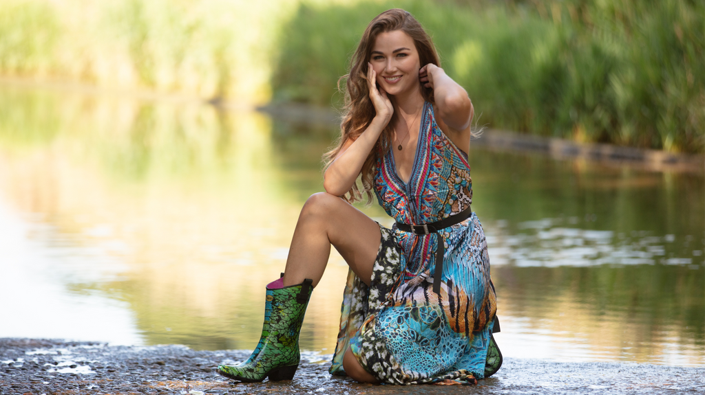 girl in bright green and blue peacock coloured cowboy boot wellies kneeling by pond in flowing kamino dress